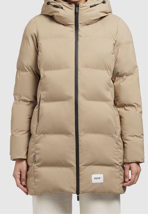 Beige gewatteerde winterjas met frontale ritssluiting, capuchon met verstelbare trekkoorden en een geborduurd logo nabij de zoom, gedragen over lichte broek.