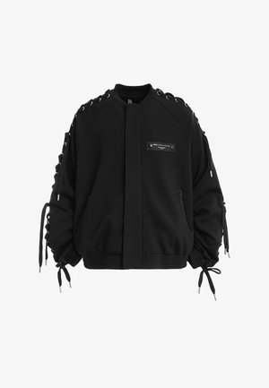 Chaqueta bomber negra con detalles de cordones en las mangas, bolsillos con cremallera, dobladillo y puños acanalados, y un pequeño parche rectangular en el pecho delantero.