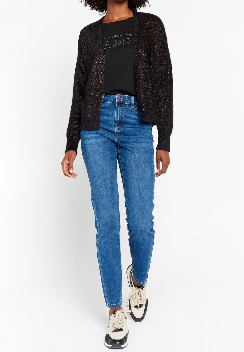 Svart texturerad cardigan med öppen front, ihopkopplad med en enfärgad svart t-shirt med metallisk tryck, blå tajta jeans och beige sneakers.