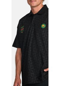 Black polo shirt with textured star pattern, featuring green and red embroidered accents and a round logo patch on the left chest.