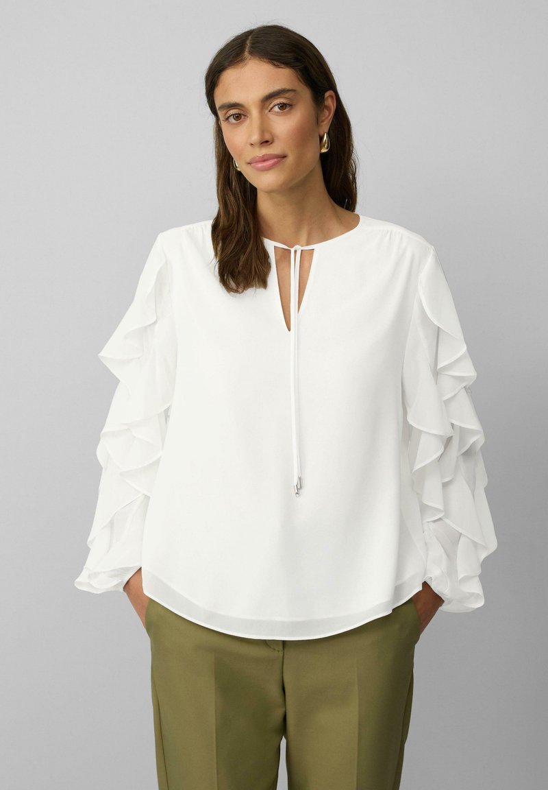 Blouse blanche avec des manches à volants, un col en forme de trou de serrure et des détails à cordon. Portée avec un pantalon ajusté vert olive sur un fond neutre.