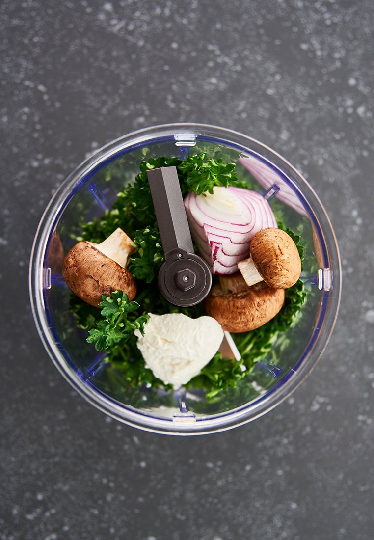 Un bol contenant du persil haché, des champignons, de l'oignon rouge et du fromage à la crème en forme de cœur, avec une lame de mixeur grise au centre.