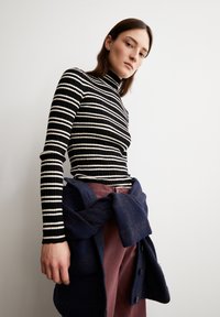 Schwarz-weißer gestreifter, gerippter Rollkragenpullover, kombiniert mit kastanienbraunen Hosen und einem dunkelblauen Pullover, der um die Taille gebunden ist. Glatte Textur, enganliegendes Design.