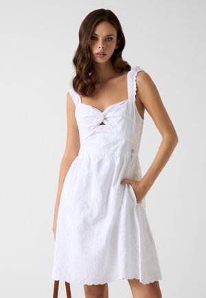 Młoda kobieta o falowanych brązowych włosach nosi białą, bezrękawnikową sukienkę z wiązanym biustem, falowanym dołem i prawą ręką w kieszeni na jednolitym tle.