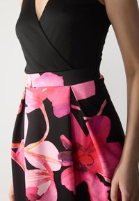 Robe noire avec un design en v et une jupe à motifs floraux rose et violet ; fabriquée dans un tissu lisse avec des détails plissés.