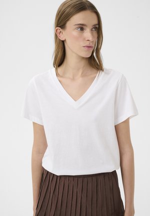 Młoda kobieta o jasnobrązowych włosach, ubrana w biały T-shirt z dekoltem w serek i brązową plisowaną spódnicę, patrząca w bok na tle jednolitego tła.