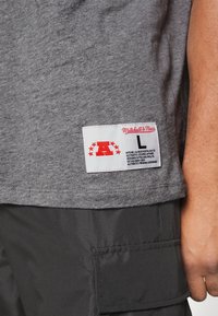 Grå bomulds t-shirt med et hvidt mærke med røde accenter. Mærket indeholder brandnavnet "Mitchell & Ness," størrelse "L" og plejeinstruktioner.