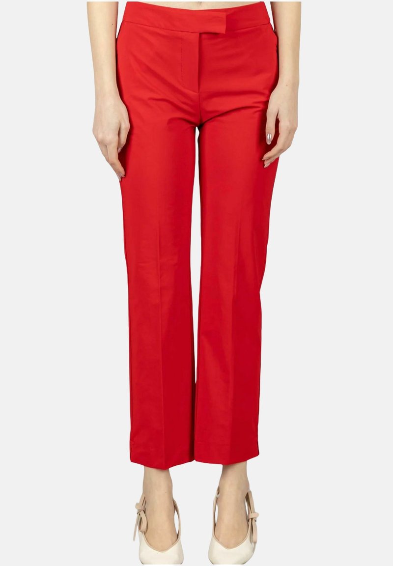 Femme portant un pantalon droit rouge vif et des chaussures blanches à talons hauts, debout devant un fond uni.