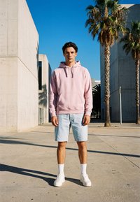 Jonge man in een roze hoodie, lichte denim shorts, witte sokken en sneakers, staand buiten bij betonnen gebouwen en palmbomen onder een heldere blauwe lucht.