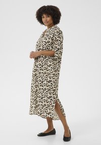 Vestido de estampado de leopardo en beige y marrón, con corte holgado, mangas de tres cuartos y aberturas laterales. Llevado con zapatos planos negros.