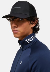 Marine blauw zip-up jack met een hoge kraag en een wit logo op de kraag. Zwarte mesh pet met "PeakPerformance" in witte tekst aan de voorkant.