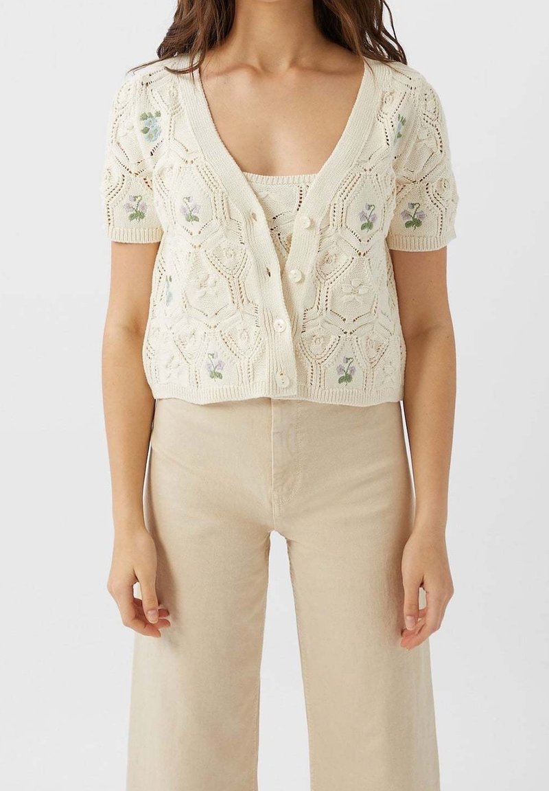 Ivory knitted cardigan with a scalloped texture, featuring floral embroidery, short sleeves, and button closure, worn over a matching top.