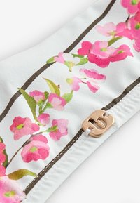 Material alb cu model floral roz, tulpini maro și o mică etichetă metalică aurie gravată cu litera „b”, atașată cu ață albă.