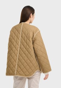 Beige gesteppte Jacke mit lockerer Passform, die ein Diamantmuster aufweist, weißen Besatz an den Nähten und weiten Ärmeln für Bewegungsfreiheit.