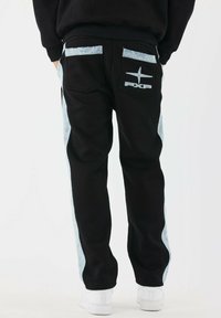 Pantalones de chándal negros con paneles laterales de denim azul, un logo bordado en los bolsillos traseros y un corte relajado. Se llevan zapatillas blancas.