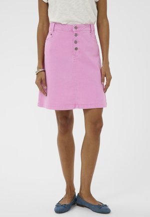 Žena v ružovej denimovej sukni s vysokým pásom so štyrmi prednými gombíkmi, v kombinácii s modrými balerínkami a bielym topom.