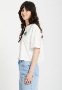 T-shirt blanc court en coton doux, avec un motif brodé vert et rouge sur la poitrine et des manches courtes.