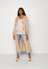 Casaco trench bege com gola oversized e botões, usado sobre uma t-shirt às riscas, combinado com jeans azuis e sandálias brancas com tiras.