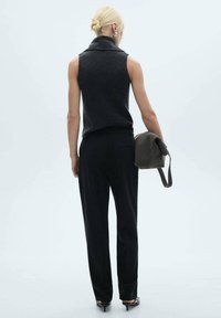 Grey sleeveless top with a high collar, paired with black trousers. A small dark bag is held in the left hand. Simple, unadorned design.