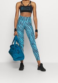 Musta urheilu rintaliivi, jossa on "Just Do It" -teksti, siniharmaaraidalliset legginsit ja teal-värinen urheilukassi. Asusteena mustat urheilukengät.