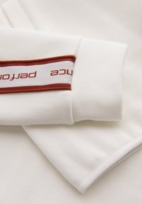 Manche blanche de sweat-shirt avec bord-côte et bande blanche bordée de marron comportant un texte marron partiellement visible.