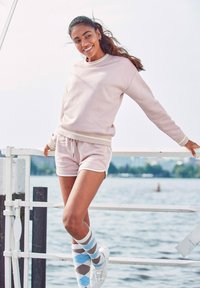 Svetloružová mikina a šortky, vyrobené z mäkkého materiálu. Biele akcenty na manžetách a vreckách. Ponožky s kosoštvorcovým vzorom v modrej, sivej a bielej farbe.