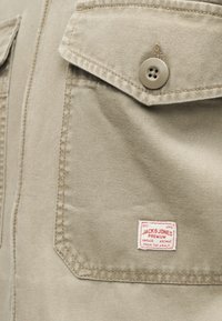 Beež kangast tasku nööpidega klapiga ja punane-valge Jack & Jones rõivamärgiga, mis on õmmeldud all paremasse nurka.