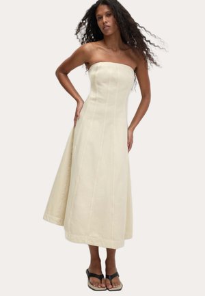 Mouwloze, crèmekleurige jurk van katoen, met een uitlopende rok, verticale naden en een zoom die tot halverwege de kuit reikt. Gemaakt in combinatie met zwarte sandalen.