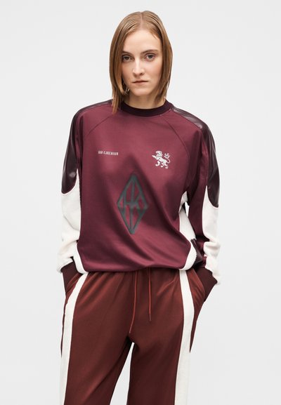 Haut de sport bordeaux avec des accents crème, arborant un emblème de lion et un logo géométrique ; assorti à un pantalon bordeaux avec des bandes latérales blanches.