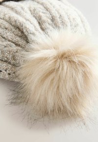 Gebreide muts van een gestructureerde beige stof met subtiele glans, afgewerkt met een grote crèmekleurige bontpompon, die fijne haartjes vertoont.