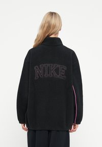 Crna flis jakna s visokim ovratnikom, koja na leđima ima hrabri "NIKE" logotip. Bočne akcenti u jarkoj ružičastoj boji. Tekstura i kroj koji je prevelik.