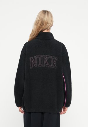 Čierna fleecová bunda s vysokým golierom, s výrazným logom "NIKE" na chrbte. Bočné akcenty v jasnej ružovej farbe. Textúrovaný, voľný strih.