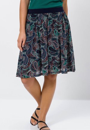 Femme portant une jupe paisley longueur genou avec des motifs bleus, verts et rouges, associée à des sandales noires à lanières et un haut vert.