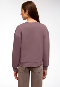 Femme aux longs cheveux bruns portant un sweat-shirt à col rond mauve et un jean gris délavé, vue de dos sur un fond blanc uni.