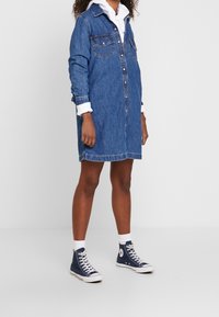 Niebieska koszulowa sukienka z denimu, z długimi rękawami, zapięciem na guziki z przodu, dwoma kieszeniami na piersi oraz prostym krojem. Połączona z niebieskimi sneakersami za kostkę.