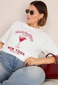 Bílé oversized tričko s červenou grafikou koktejlové sklenice a textem "Manhattan Est. 1870 New York Social Club." Pohodlné denimové džíny a burgundská kabelka.