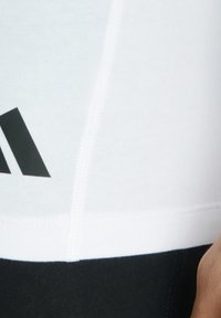 Bílý sportovní top s černým logem, hladká tkanina a přiléhavý střih. Viditelné švy podél okrajů a v pase.