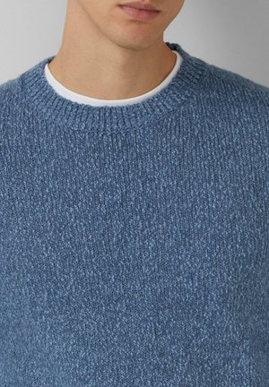 Blauer gestrickter Pullover mit geripptem Rundhalsausschnitt, aus strukturiertem Stoff und mit lockerer Passform, über einem weißen Hemd getragen.