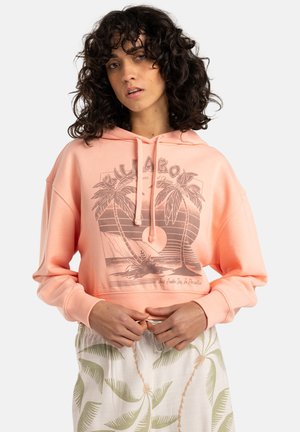 Cropped hoodie rosa chiaro realizzato in un materiale morbido, con una grafica di palma e cappuccio con cordino. Abbinato a pantaloni chiari con fantasia.