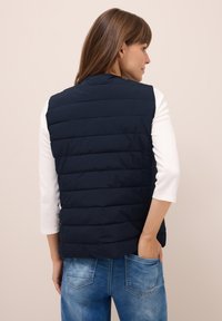 Gilet imbottito blu con texture trapuntata, colletto alto e tasche laterali, indossato sopra una maglietta bianca e jeans blu, che mostra una finitura di tessuto liscia.
