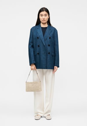 Woman wearing dark blue double-breasted blazer, black top, white wide-leg pants, beige shoes, holding beige patterned handbag by side.