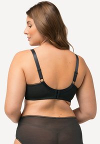 Soutien-gorge noir avec une texture de tissu lisse, des bretelles réglables et une fermeture à crochets au dos, présentant des bords festonnés.