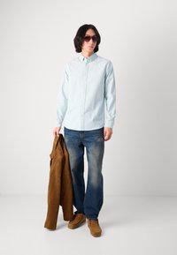 Light blue striped shirt, blue jeans, brown suede jacket, and brown shoes. Model wearing sunglasses; standing with jacket draped over arm.