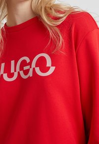 Sudadera roja con un gran logo blanco con letras curvas. La tela parece suave, con un cuello redondo y un corte holgado.
