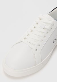 White leather sneaker with flat laces, rounded toe, subtle logo detail, and textured side panels. Smooth finish with cushioning on the collar.