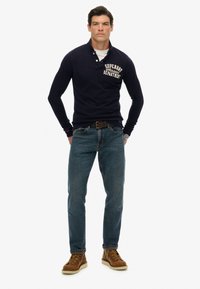 Camisa polo de manga larga en color navy con letras blancas, combinada con jeans azules y botas de cordones marrones. Presenta un cuello y un diseño ajustado.