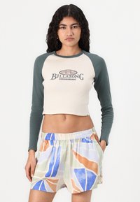 Top corto color beige con maniche raglan verdi, caratterizzato da un logo "Billabong". Pantaloni corti a fantasia floreale nei colori pastello con una cintura elastica.