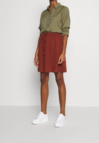 Camisa a rayas verde oliva con mangas remangadas, combinada con una falda color óxido con cintura ajustable. Lleva zapatillas blancas.