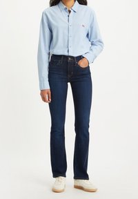 Chemise bleu clair à manches longues, jeans bleu foncé et baskets blanches. La chemise présente un petit logo rouge ; les jeans ont des poches classiques.