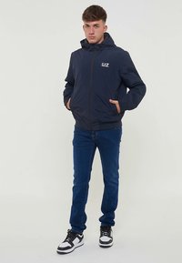 Tamsiai mėlyna striukė su gaubtu, priekyje su užtrauktuku ir EA7 logotipu ant krūtinės. Derinama su mėlynomis džinsais ir juodai baltais sportbačiais.